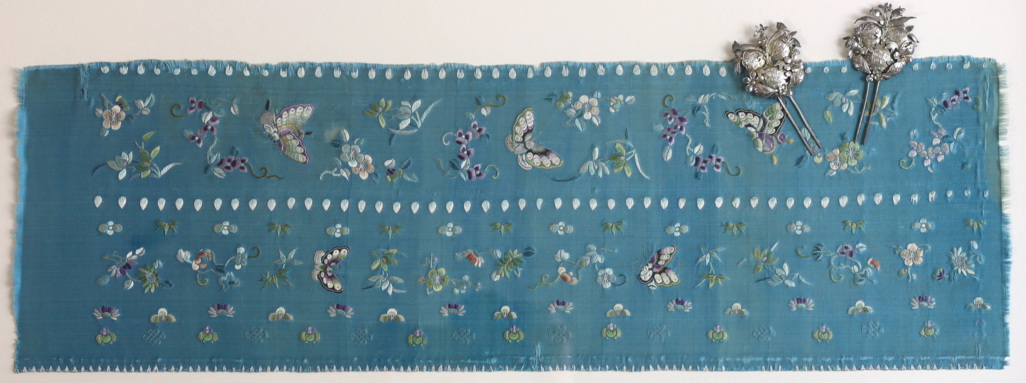 A pair of late 19th century Chinese blue silk multi-coloured embroidered panels, possibly a pair of wide sleeve bands to a robe, each mounted with two white metal ornaments, 19cm wide, 61cm high